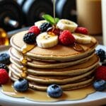 Stack of fluffy protein pancakes topped with fresh fruit and honey, served on a white plate with a protein shaker in the background.