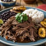 Authentic ropa vieja recipe served with rice, black beans, and plantains on a rustic Cuban dining table.